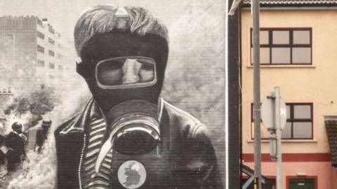 A painting of a mural of a young boy in a gas mask in the Creggan area of Derry. It is a black and white mural. He is wearing a dark jacket, a striped shirt and a pin depicting the island of Ireland.