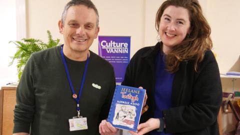 Adrian Cain, who has short greying hair and is wearing a black jumper, and Ruth Keggin-Gell, who has long brown hair and is wearing a black jacket over a blue top, holding a copy of the book, which has a blue cover with a black and white illustration. There is a purple Culture Vannin banner in the background.