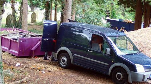 CCTV image of a blue van, with a man at the back of it behind its open doors. You can just see the man's legs and a purple large skip with waste in it. In the distance are gravestones and trees.