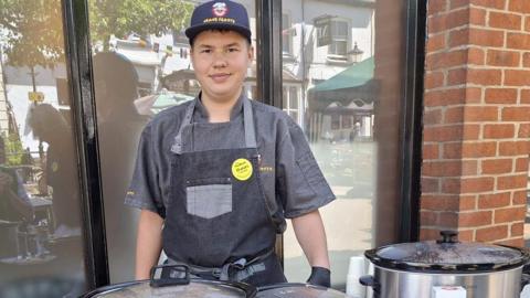 Dexter stood in his all grey chef uniform stood in front of slow cookers on a table with recipe cards, spices, tins and flatbreads on the table.