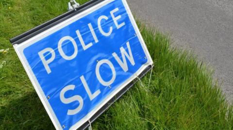 A stock picture of a blue sign with white writing reading "Police Slow" on a grass verge beside a road