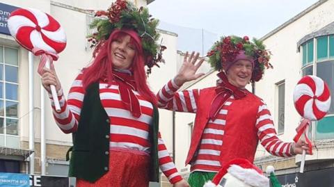 Two smiling stilt walkers - a man and woman - posing for the camera. They are dressed as elves in red and white stripey jumpers and carrying giant lollypops.