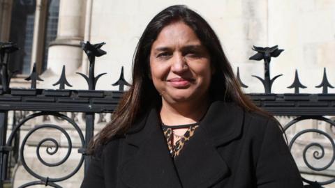 Seema Misra standing in front of some black railings, looking directly at the camera with a half smile. She is wearing a black coat. 