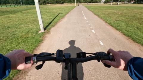 A first-person view of someone riding a bicycle along a paved path through a flat grassy landscape in a park. The rider’s hands can be seen on the handlebars, and their shadow stretches ahead on the path. The path itself has dashed white lines down the middle.