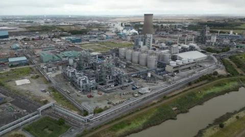 An industrial site with large silos, cooling towers and chimneys. Pipework is also visible stretching around the site. The image has been captured from a drone and shows the size and scale of the operation. Green fields in the surrounding area are visible and stretch into the distance.