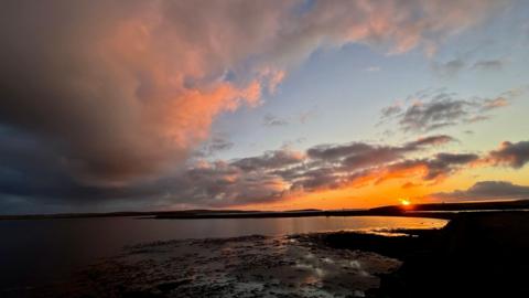 Partly cloudy sky over the coast with sun setting in the distance.