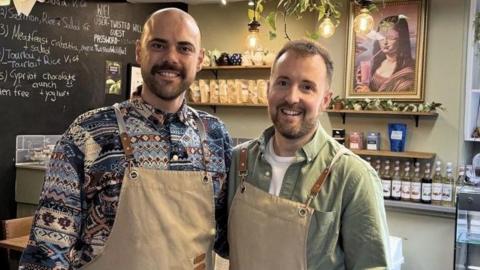 Constantinos Theophanous and Scott Thain stand together in a cafe. They are wearing brown aprons and are both smiling. There is a black board with drinks and food listed on it and to the right of them there are several syrups and coffees and teas. Constantinos has no hair and is wearing a patterned blue shirt while Scott has short brown hair and is wearing a green shirt.