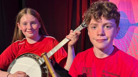 A young boy and girl wearing red t-shirts. They are both smiling at the camera. The girl, who has long fair hair is playing a banjo. The boy, who has short curly brown hair is holding pipes. 