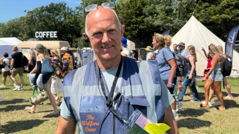 Steve Wood smiles as he poses for a photograph on a sunny day with Isle of Wight festival-goers passing behind him on the festival site. He wears a vest jacket with the words The Welfare Crew on it. He is bald with short, white hair at the sides and glasses pushed back over his head.