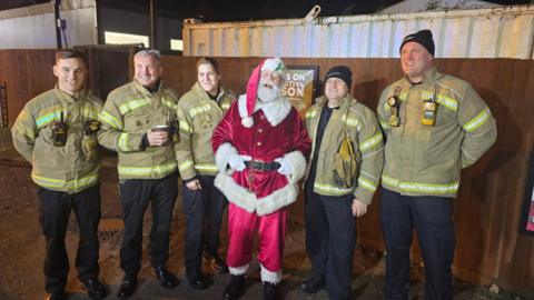 Five firefighters standing next to a man dressed as Santa outside. They are standing in front of a wooden fence which has a shipping container behind it. 