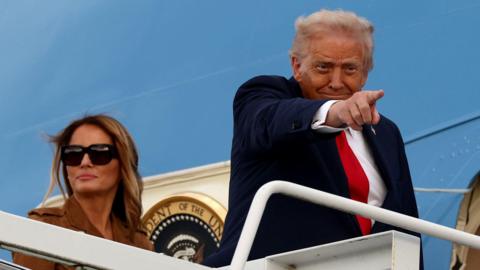 Donald Trump is wearing a navy suit jacket, white shirt and red tie. He is standing at the top of a staircase leading to the blue Air Force One jet. He is pointing towards someone on the ground. Behind him, Melania Trump wears a brown jacket and dark sunglasses.