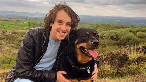 Oliver Robinson with medium length brown hair, wearing a dark jacket and light blue tshirt smiles at the camera with his arms around a black and tan rottweiler dog with countryside and fields in the background