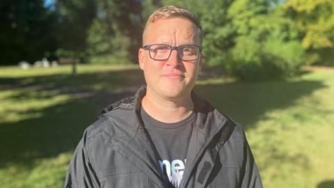 Kieran, looking at the camera, he is standing by a green area, wearing glasses and dark clothing