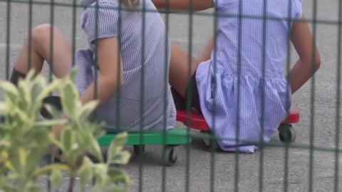 The image shows two individuals sitting on small wheeled boards, 
one green and one red, on a paved surface. Both are dressed in similar checkered clothing. The photo is taken through a fence, with some foliage visible in the foreground.