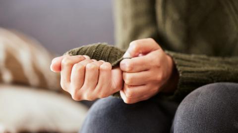 A woman sits on a sofa. She is pulling on a sleeve of her green jumper in a sign of feeling anxious.