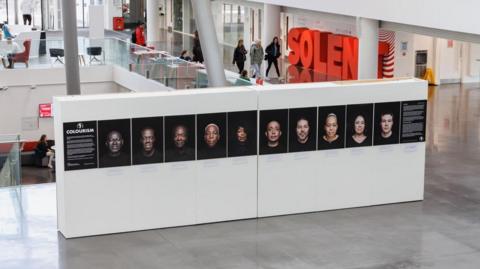 A giant wide white plinth with 10 headshot images is on display inside a building with a mezzanine floor visible in the background with people walking past.