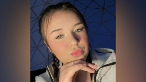Toya Jefferies wearing a grey hoodie and looking at the camera. She has her right hand under her chin and she is pouting. The background is purple.