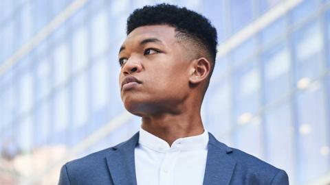 Randall Goosby - head and shoulders shot of a young black man in a blue jacket and white shirt. He is looking to the left.