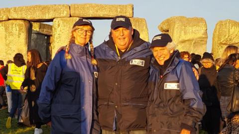 Three members of the Salisbury Street Pastors in blue and black coats in front of Stonehenge