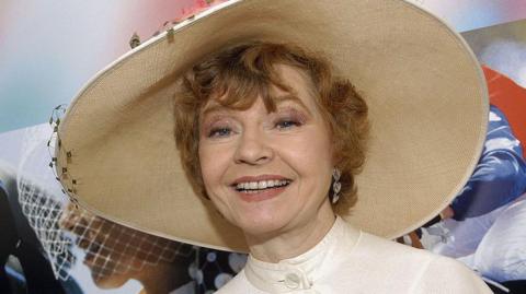 Prunella Scales is shown in middle age wearing a prominent wide-brimmed hat and smiling.