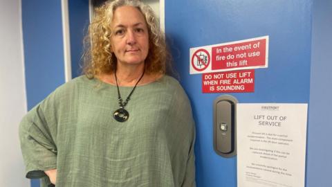 A woman with curly light hair and dressed in green, stands with her hand on her hip next to a sign that says, 'lift out of service'