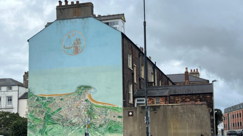 A large mural painted on the end wall of a tall terraced building at a street corner. The mural depicts an aerial view of a coastal town, including streets, housing estates, green open spaces and a sweeping sandy bay that curves around the coastline. The artwork is painted in soft shades of green, beige, blue and brown, giving it the appearance of a detailed map or landscape illustration. Near the top of the wall, above the map, there is a circular emblem containing a stylised image of a sailing ship beside a tower or castle-like structure. The emblem is outlined in a pale golden colour and stands out against the light blue background of the upper wall.
The building itself has a chimney stack at the top and forms the end of a row of dark-brick terraced houses that extend into the background on the right. Adjacent to the mural is a plain grey wall, which is much lower than the painted building.