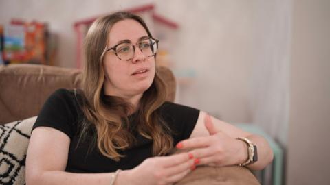 Katy, who wears glasses and a black T-shirt, talks to BBC Panorama from her sofa. Toys can be seen in the background.