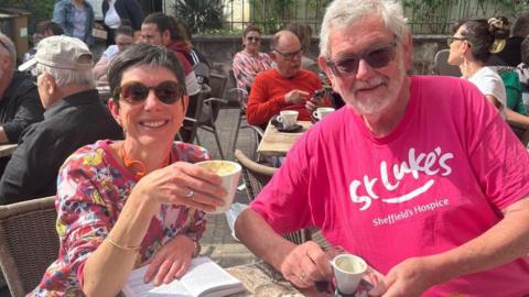 A white haired man in a bright pink t-shirt sits with a younger lady with dark short hair in an outdoor cafe.