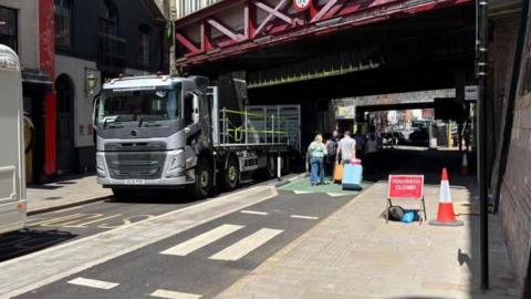 There is a road with a floating island in the middle. On the path, there is a red street sign which says 'Footpath closed,' and a traffic cone. On the one side, there is heavy traffic, including a big Volvo lorry. In the other lane, there is a cycle line which is being used by a family pulling suitcases.