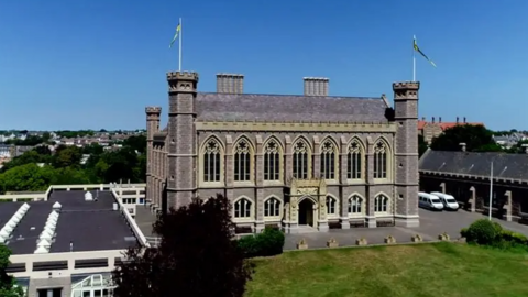 An aerial view of Victoria College - an all boys school in Jersey which looks like a castle. Field in front and blue skies behind.