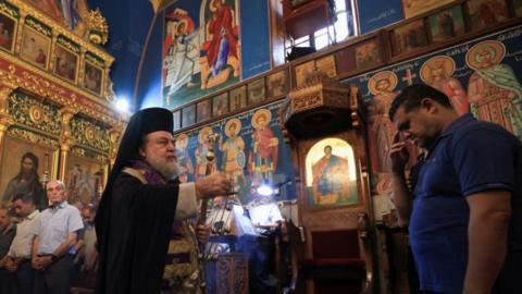 A priest stands in the Holy Family Church in Gaza as a mourner receives blessings during a funeral for those killed in the strike - 17 July 2025