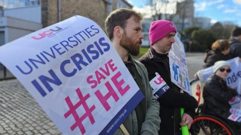 A protester with a beard holds a banner saying 'universities in crisis' with two other protesters, one in a wheelchair, in the background