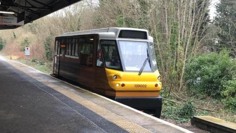 A small maroon colours shuttle train with a yellow front. It is a single carriage, and has stopped in front of a platform