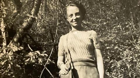 A black and white picture of Alice Moody. She is smiling at the camera.  Alice is wearing a jumper and skirt and she is holding a handbag.  Trees and bushes are in the background.