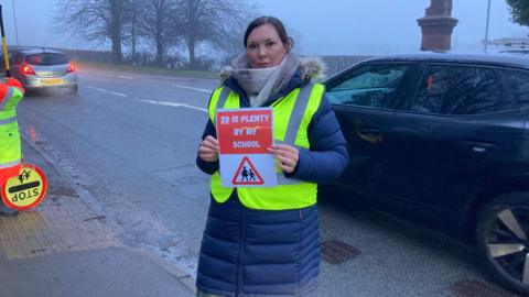 A woman wearing a long winter coat and a hi-vis jacket, stands at the side of the road and holds a road safety sign which says "20 is plenty by my school".