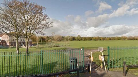 A large playing field behind a green metal railing with a row of trees in the distance.