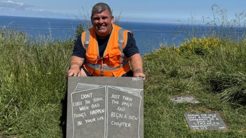 A man wearing an orange Hi-Viz bib and holding a slate sign next to a cliff edge