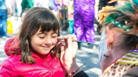 A girl in a pink coat has her face painted at a Purim festival