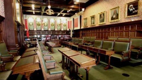 An empty House of Keys chamber, with wooden seating and benches upholstered with avocado green leather with a matching carpet. The chamber has wooden panelled walls and portraits of previous Speakers of the House of Keys on the righthand side and stained glass windows at the far end.