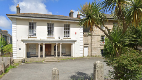 The Redruth Albany RFC clubhouse is a white two storey Georgian style granite house with three long windows on the upper floor. There are two columns each side of the wooden entrance door and a chimney on each side of the roof. A tarmac car park is empty in front of it which is outlined with carved granite pillars. There are palm trees in the hedge lining the car park. Attached to the club house is a three storey granite building.