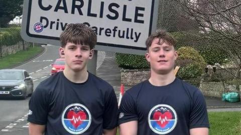 Oliver and Theo standing in front of a sign that says "Carlisle, Drive Carefully". There is a grey car in the background driving on the road. The pair are standing to the side of the road on grass. They are both wearing dark blue tops with a circle symbol on them and a red heart inside. They both have dark brown hair.
