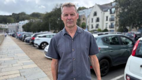 A man looking at the camera. He has very light brown hair. He is wearing a dark short sleeved shirt. He is stood in front of a row of parked cars.