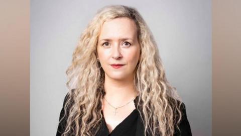 Victoria Bell is wearing a black top, belt and jacket and has long blonde curly hair. She is looking straight at the camera.