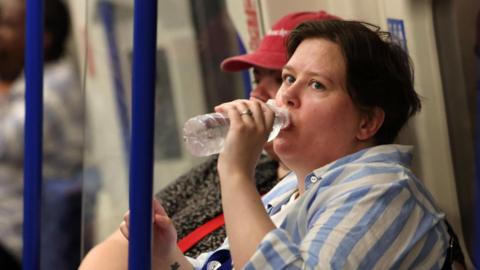 Woman in a light blue and white striped shirt sits in seats on the Tube. She drinks water from a clear bottle, her face looking slightly flushed from the heat.