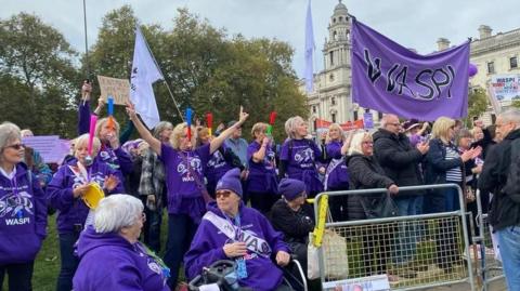 A large group of women in purple t-shirts and jumpers standing outside Parliament with banners and signs.