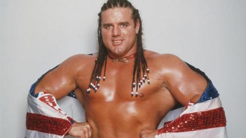 Wrestler British Bulldog stands smiling with his hands on his hips and flexing his arms. He has long, braided hair and has a sparkly red, white and blue flag wrapped around him. 
