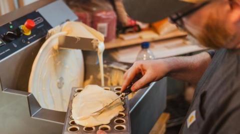A man with glasses and short beard spreads a layer of white chocolate over a tray full of cup-shaped chocolates, with a silver chocolate-making machine in the background, which features a large white wheel from which molten chocolate is dripping.