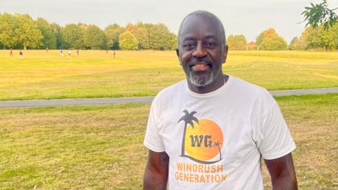 A man with short grey hair and goatee beard stands in a park. His white tshirt has a sun, palm tree and the letters WG forming a logo. Beneath the logo it says WINDRUSH GENERATION. There are trees and people playing cricket in the background on a sunny day.