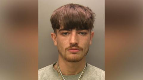 A mugshot of Noah Hetherington. He has brown hair that is longer on the top and shaved at the sides. He has blue eyes and a goatee and is wearing a grey jumper with two necklaces - a golden chain and one with white beads. He is looking at the camera with a serious expression.