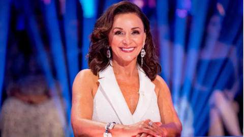 Shirley Ballas, wearing a white top, smiles from behind the judge's desk on Strictly